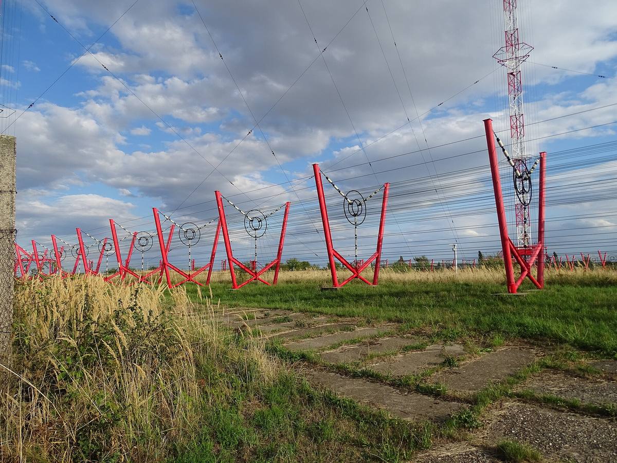 Červené ocelové stojany s izolátory pod spletí drátů, vpravo v pozadí červenobílý příhradový stožár