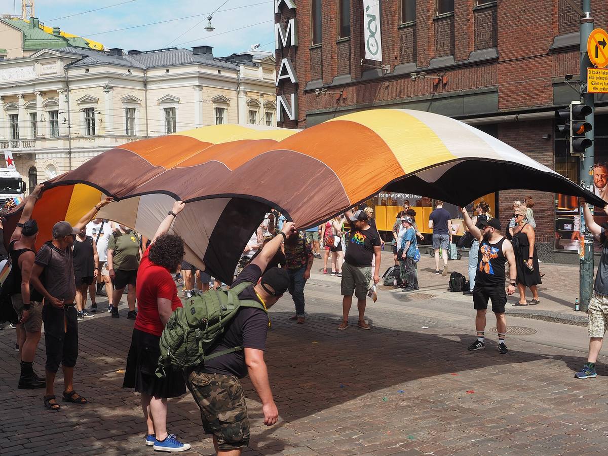 Účastníci průvodu Helsinki Pride 2022 zvedají nad hlavy velkou vlajku medvědí subkultury v hnědých, oranžových, žlutých a bílých pruzích