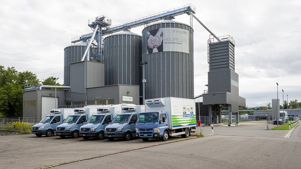Kovová sila na dřevěné pelety s nápisem Holzpellets, před nimi zaparkované dodávky