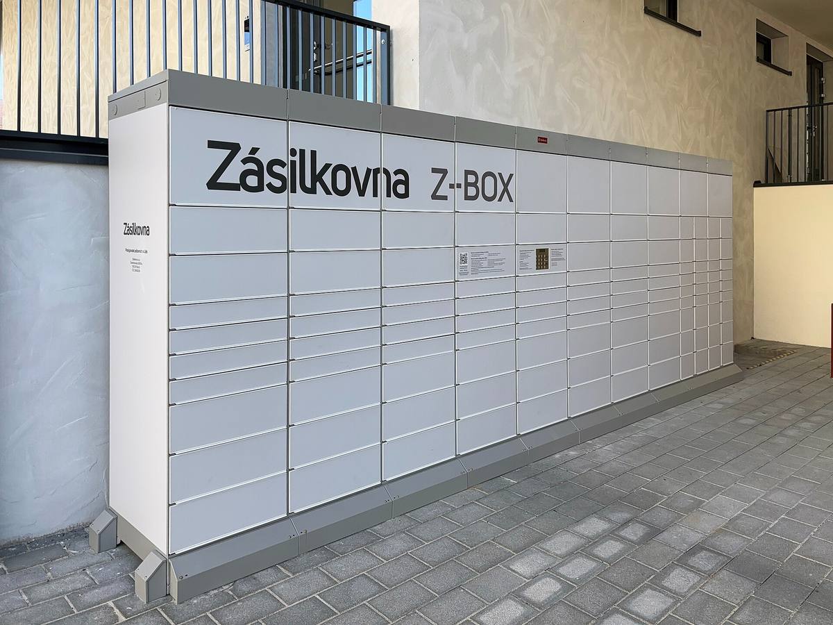 Dlouhá řada bílých schránek s nápisem Zásilkovna Z-BOX u zdi bytového domu