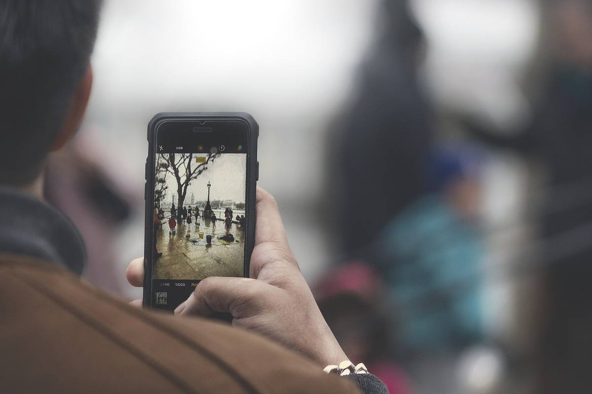 Člověk drží telefon a fotí jím nábřeží s lampou a stromem